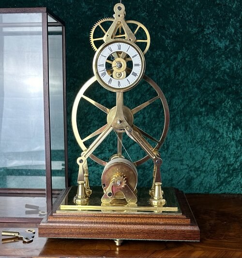 Front view of the brass great wheel skeleton clock on wooden base alongside its glass dust cover and winding key