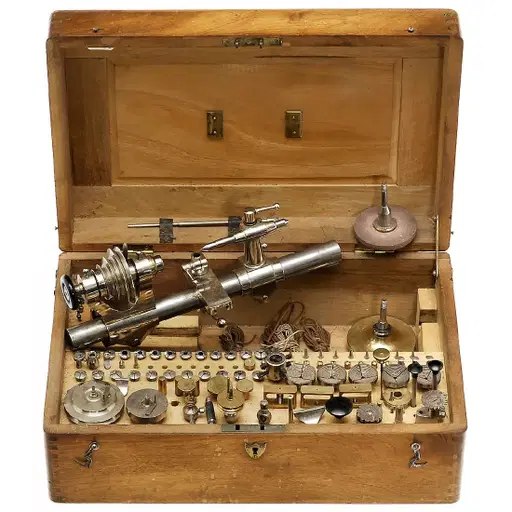 Open wooden case containing the watchmaker's lathe assembly, collets, chucks, and associated tooling
