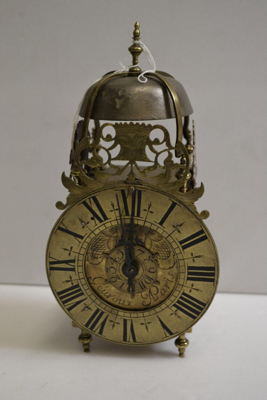 Front view of the brass lantern clock showing the dial, bell, and foliate strapwork.