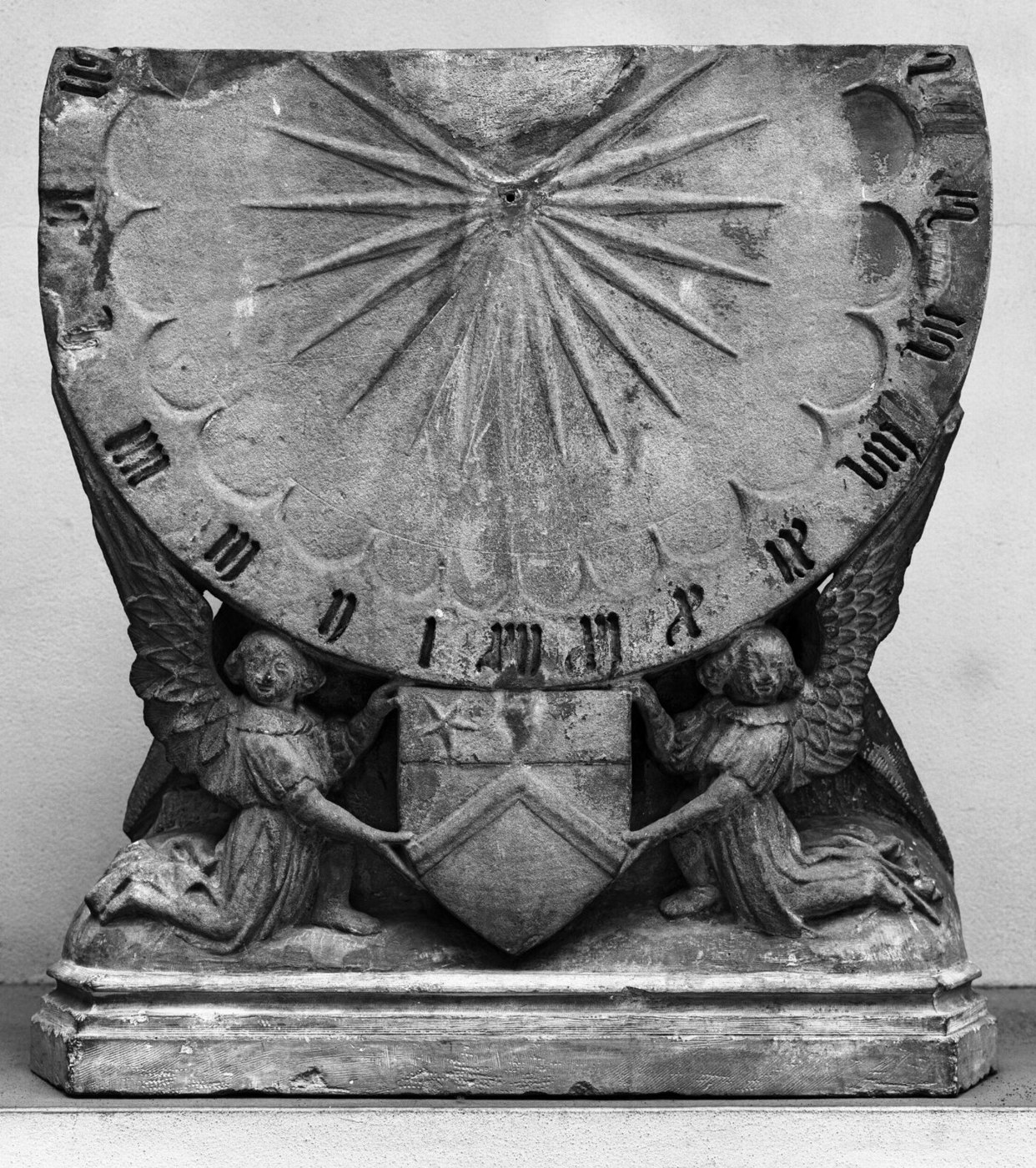 The sundial is carved from stone and supported by two kneeling angels holding a heraldic shield.