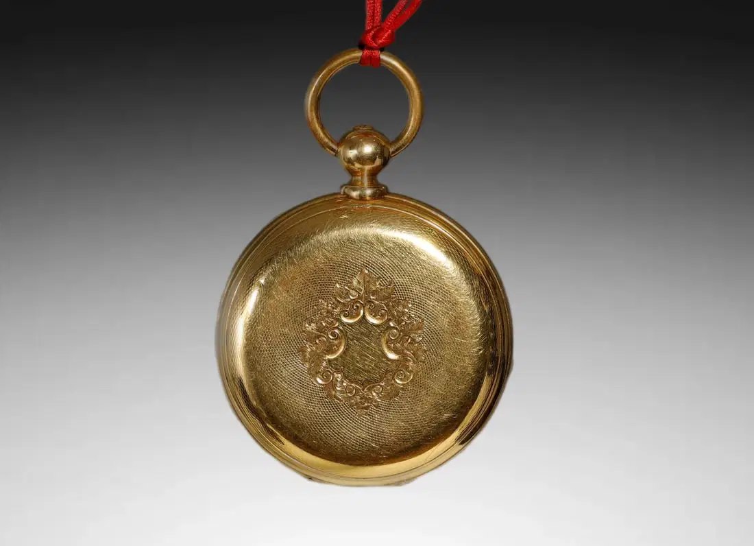 Back cover view of the closed gold hunter-case pocket watch showing the engine-turned guilloché, central cartouche, bow, and red cord