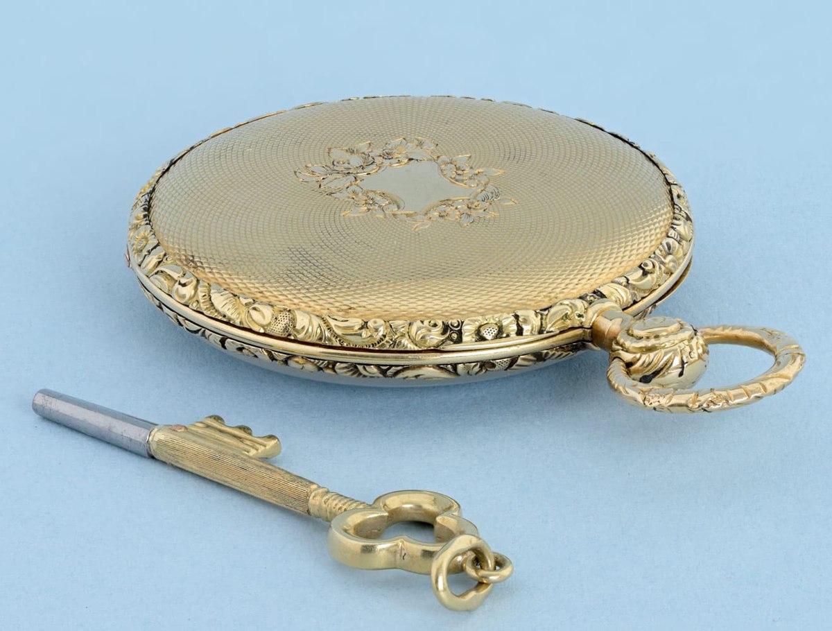 Rear view of an engine-turned gold pocket watch case showing chased bezels and an accompanying mortice-form key