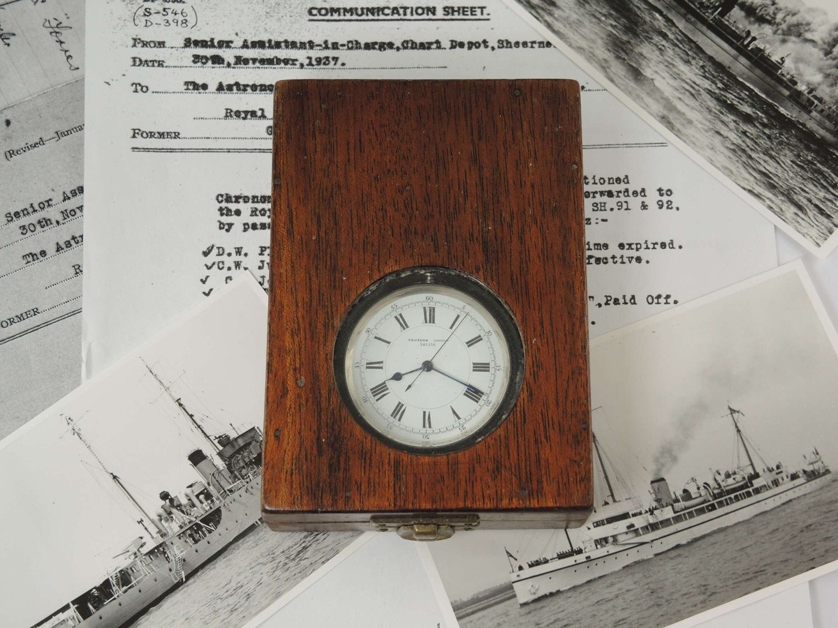 Dial of the Cousens deck watch viewed through the glazed aperture of its mahogany box atop naval service documentation