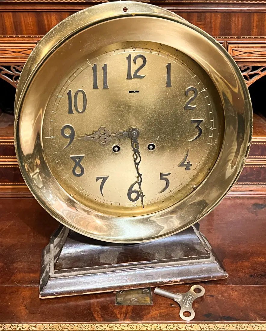 Front view of the 8-inch brass ship's bell clock mounted on a wooden stand with winding key