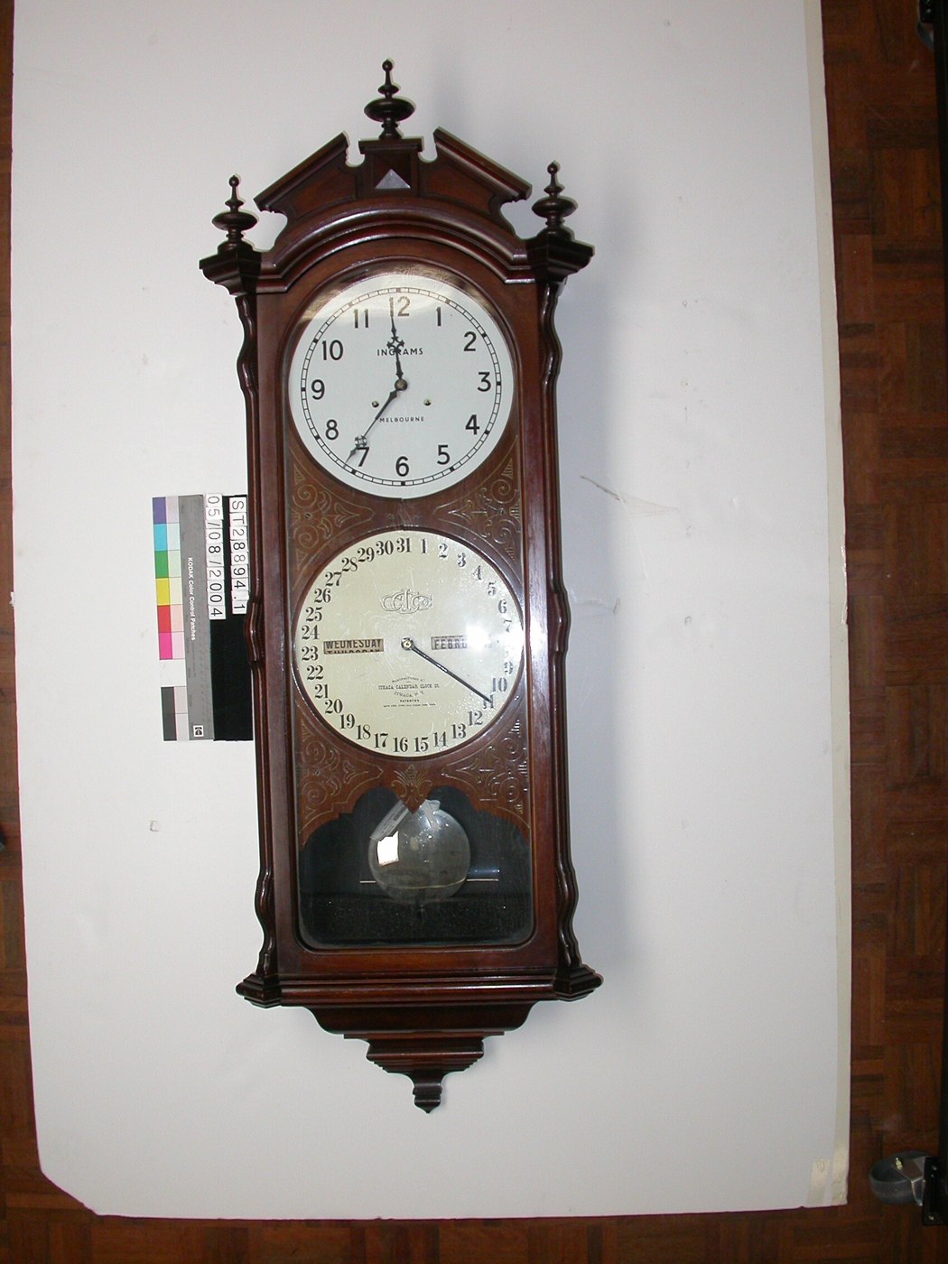 Full view of the Ithaca calendar clock featuring two dials and a pendulum aperture.