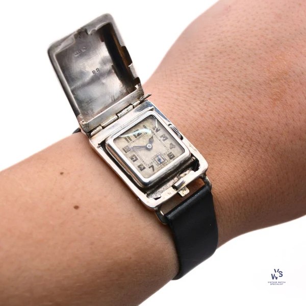 Front view of the wristwatch worn on wrist with the hinged silver outer cover opened to display the dial
