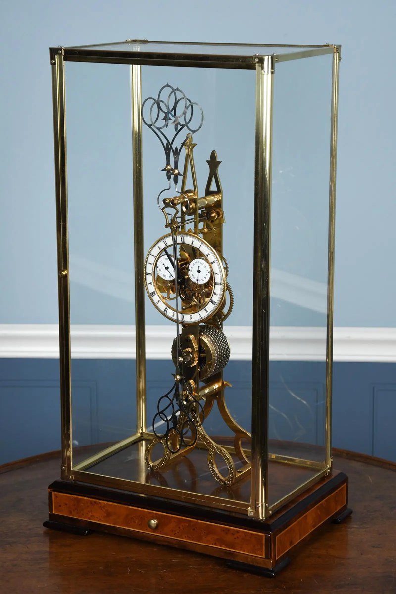 Three-quarter front view of the scissor pendulum fusee skeleton clock inside a brass-framed glass vitrine
