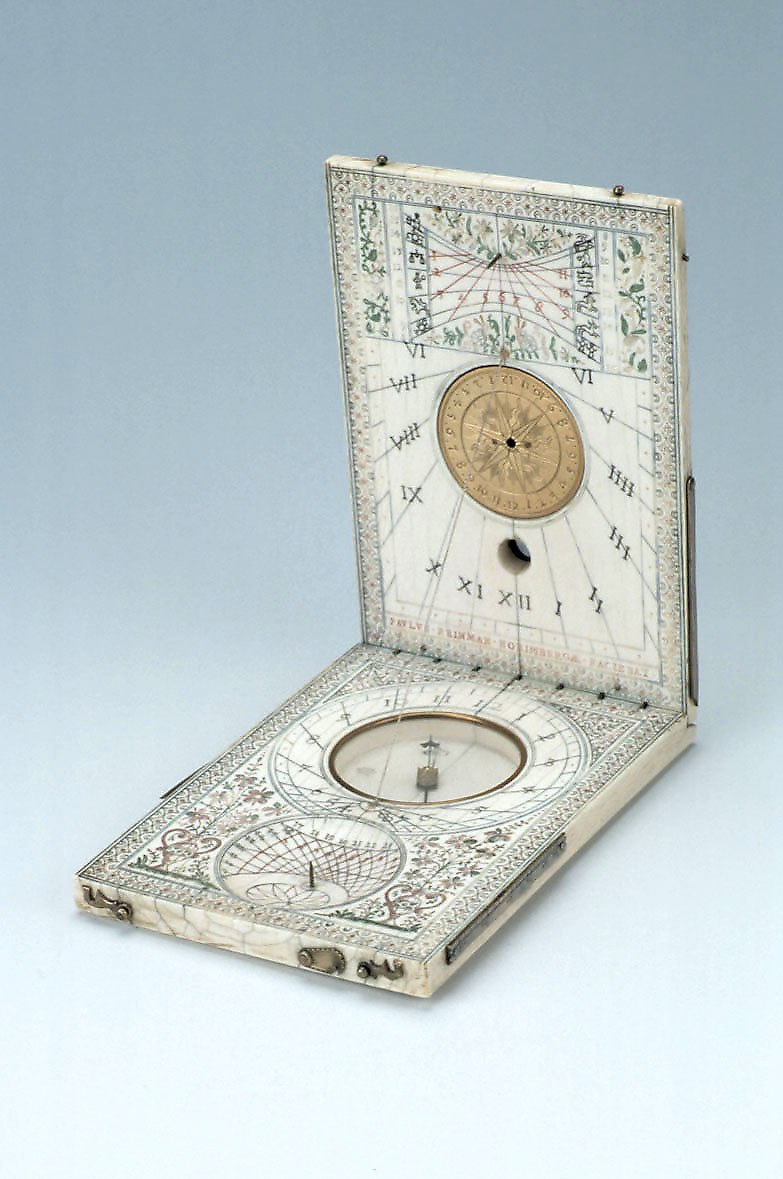An open diptych dial in ivory and brass, featuring various hour scales, a central compass, and ornate floral decorations.