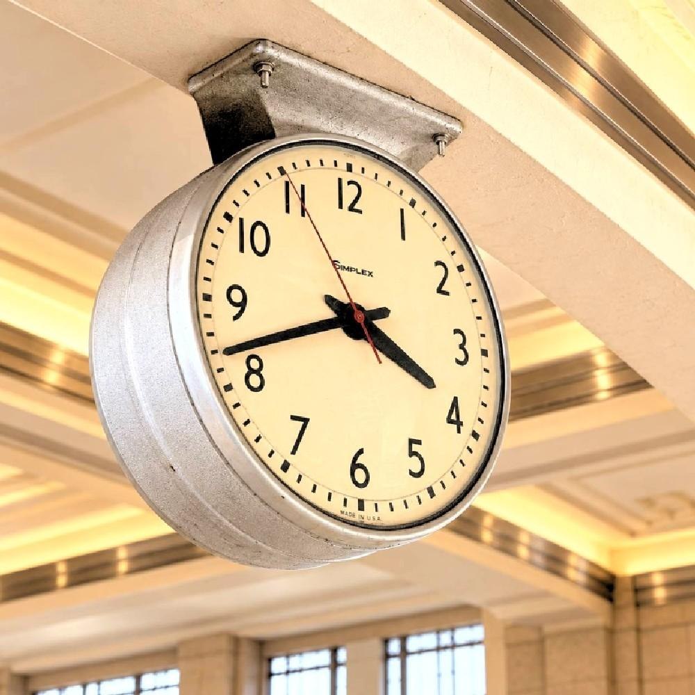 Simplex double-face waiting room ceiling clock viewed from below, showing one of the cream dials, black numerals, and the suspension bracket.