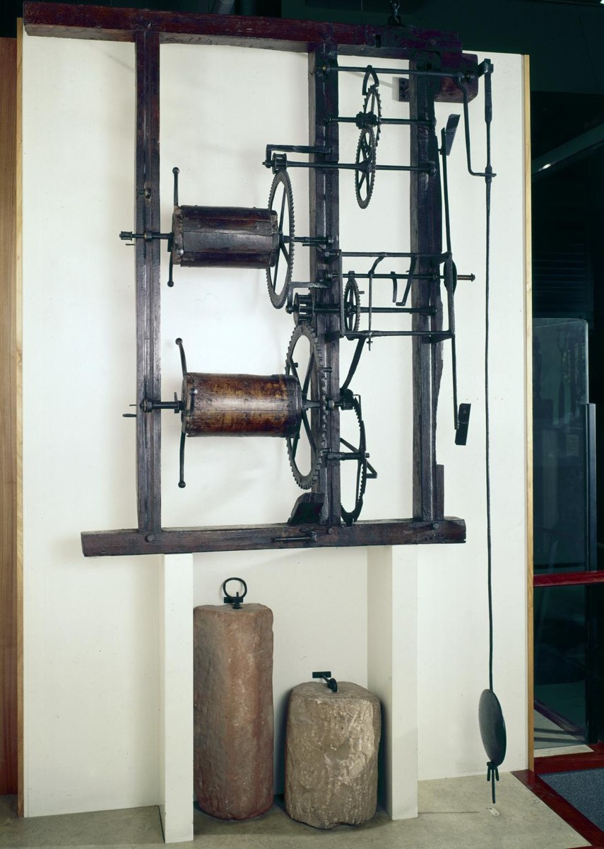 Full view of the iron turret clock mounted in its wooden frame with stone weights and pendulum