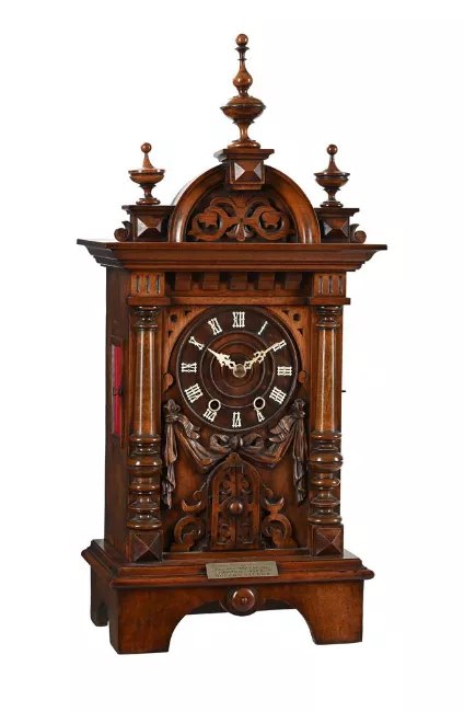 Front full view of the carved walnut trumpeter mantel clock showing the dial, finials, and base plaque