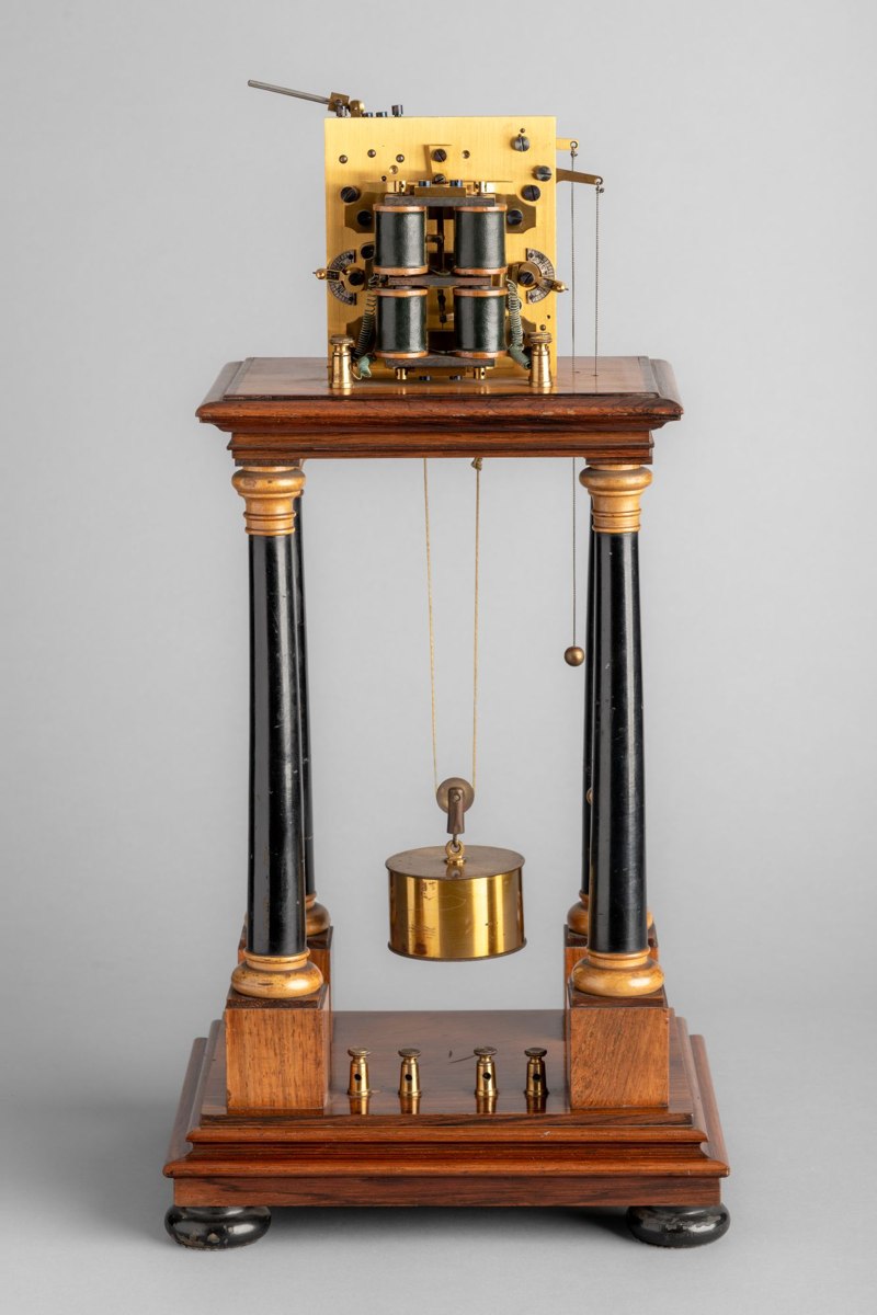 Rear view of Hipp chronoscope no. 17757 showing the pillar stand, suspended cylindrical brass weight, and movement plate with electromagnetic coils