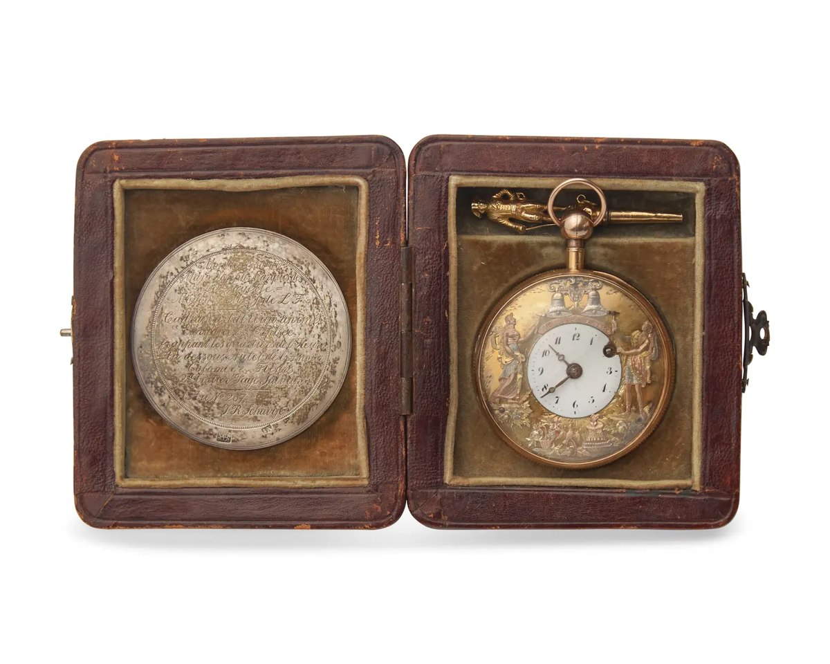 Front view of the open-face automaton pocket watch displayed inside its fitted leather case beside the engraved silver medallion and figural winding key