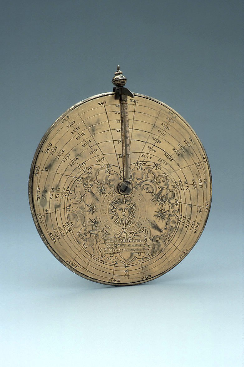 Front view of the brass Vertical Disc Dial showing the sun effigy, zodiac symbols, and the graduated gnomon rule.