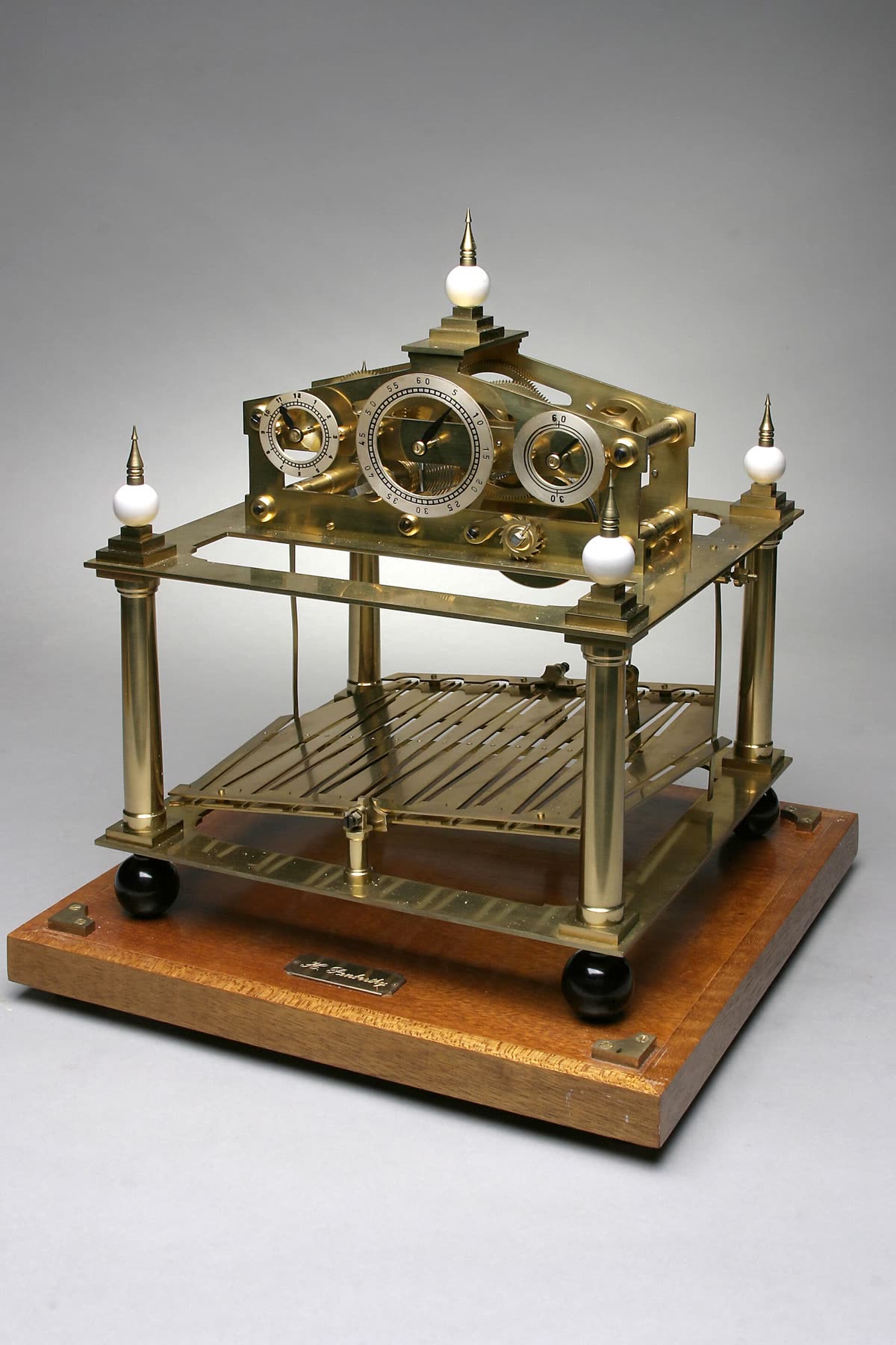 Three-quarter view of the brass Congreve rolling ball clock mechanism mounted on a wooden plinth