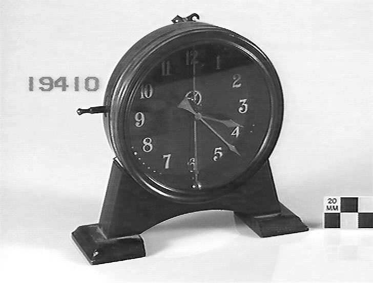 Gravity mantel clock showing the circular case on a wooden base with the manual lever on the left.